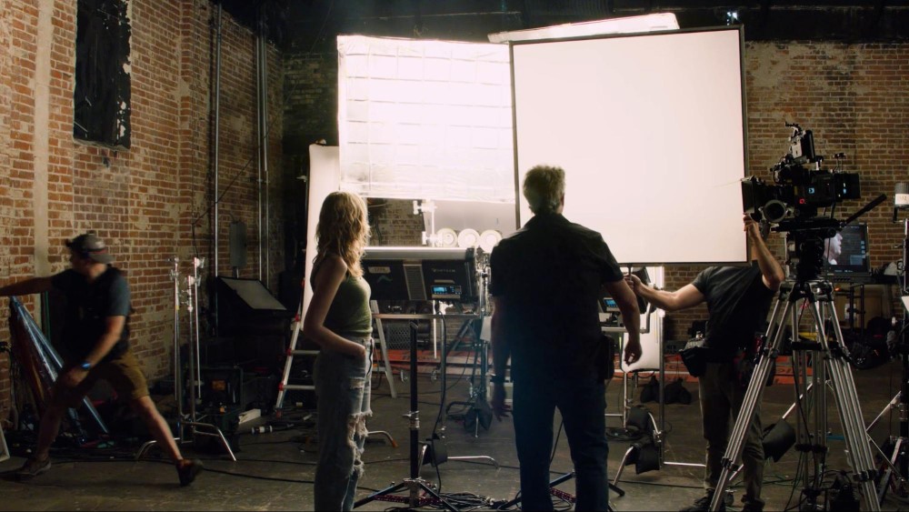 People working on a film or TV set with cameras and lighting equipment, in a brick-walled studio.