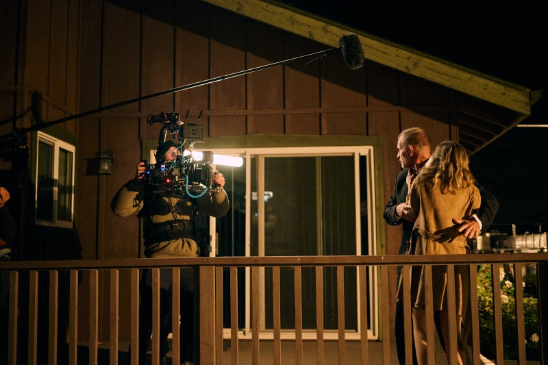 Filming crew with camera and boom microphone filming two people on a balcony at night, wooden house in background.
