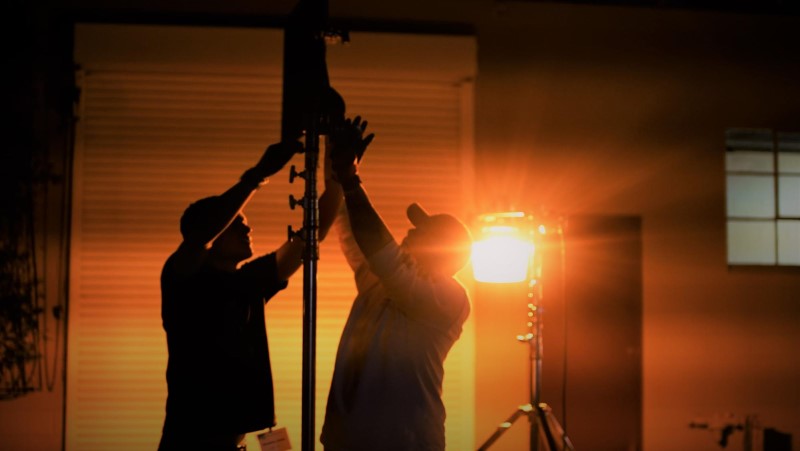 Two people hold a tall object against a bright sunset, with a light source nearby illuminating the scene.