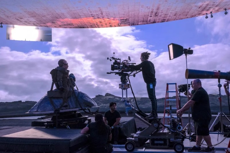 Film crew setting up camera equipment outdoors with cloudy sky and mountains in background.