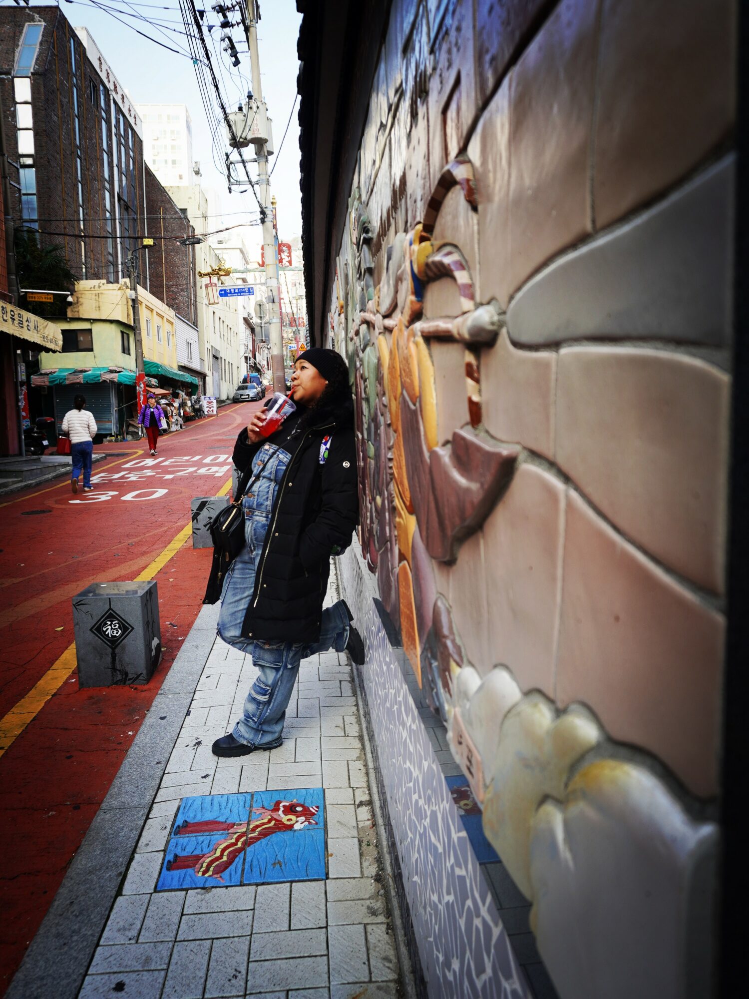 Woman standing on sidewalk near colorful mural, street scene with buildings and pedestrians in background.