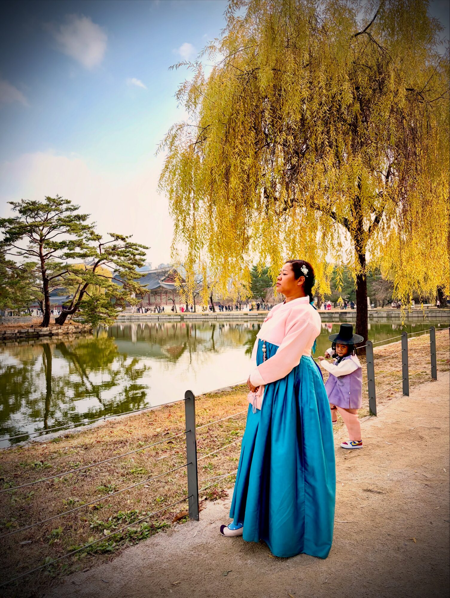 Woman in traditional dress standing near a lake with trees and a child in the background.