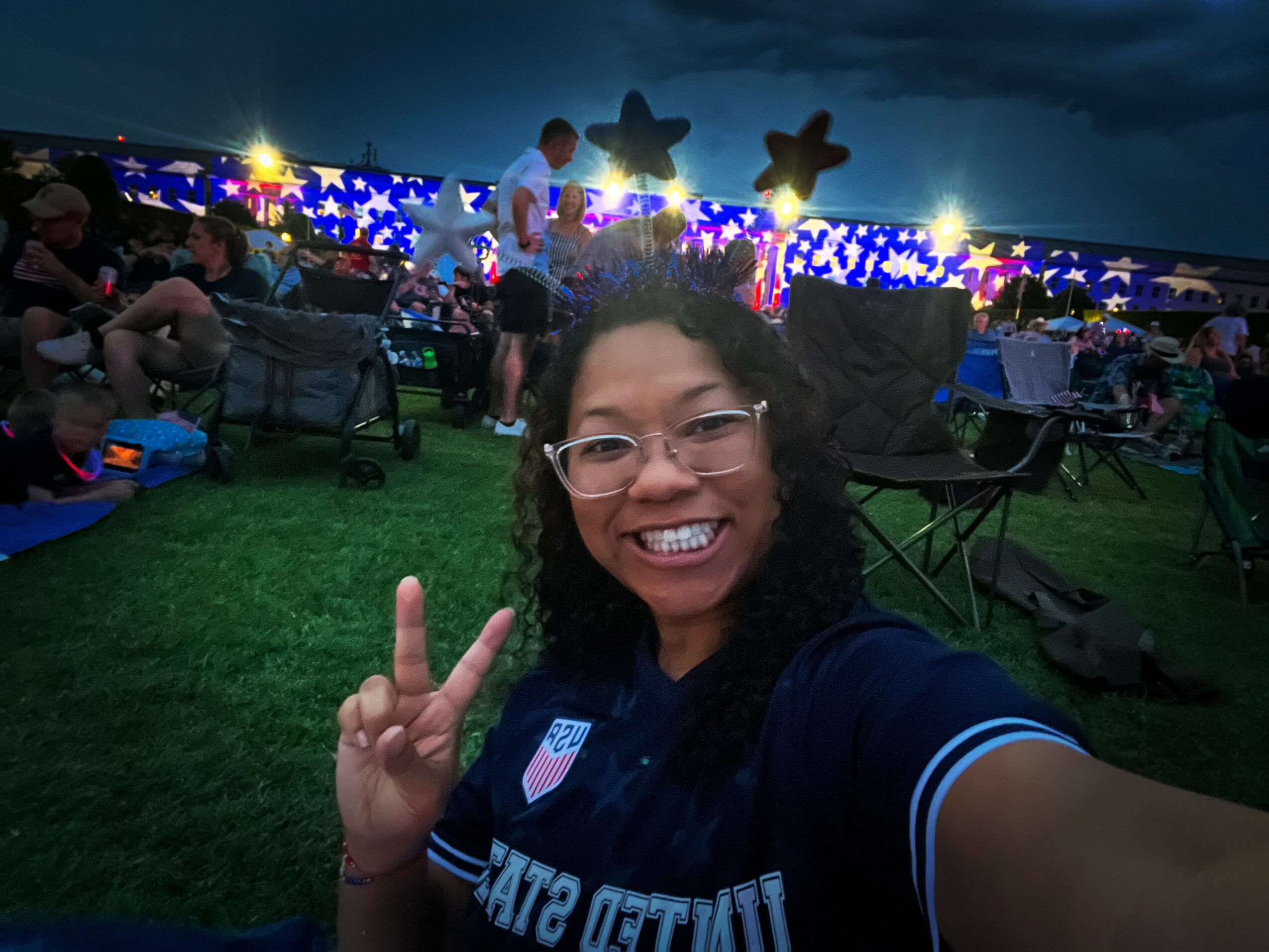 Person smiling and making a peace sign at an outdoor event during evening, with balloons and lights in background.