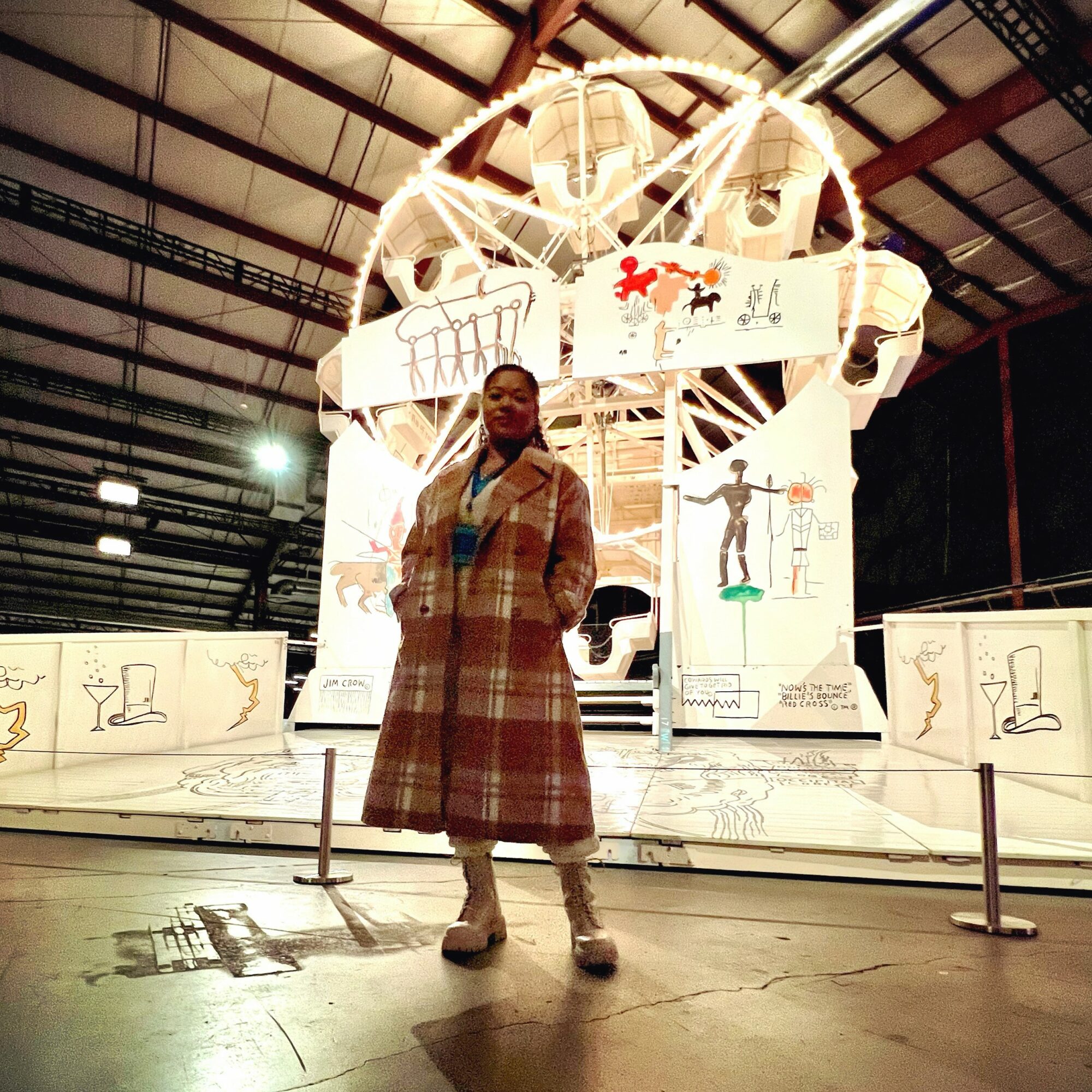Person standing in front of a large, illuminated structure inside a spacious building, with drawings and diagrams on the structure.