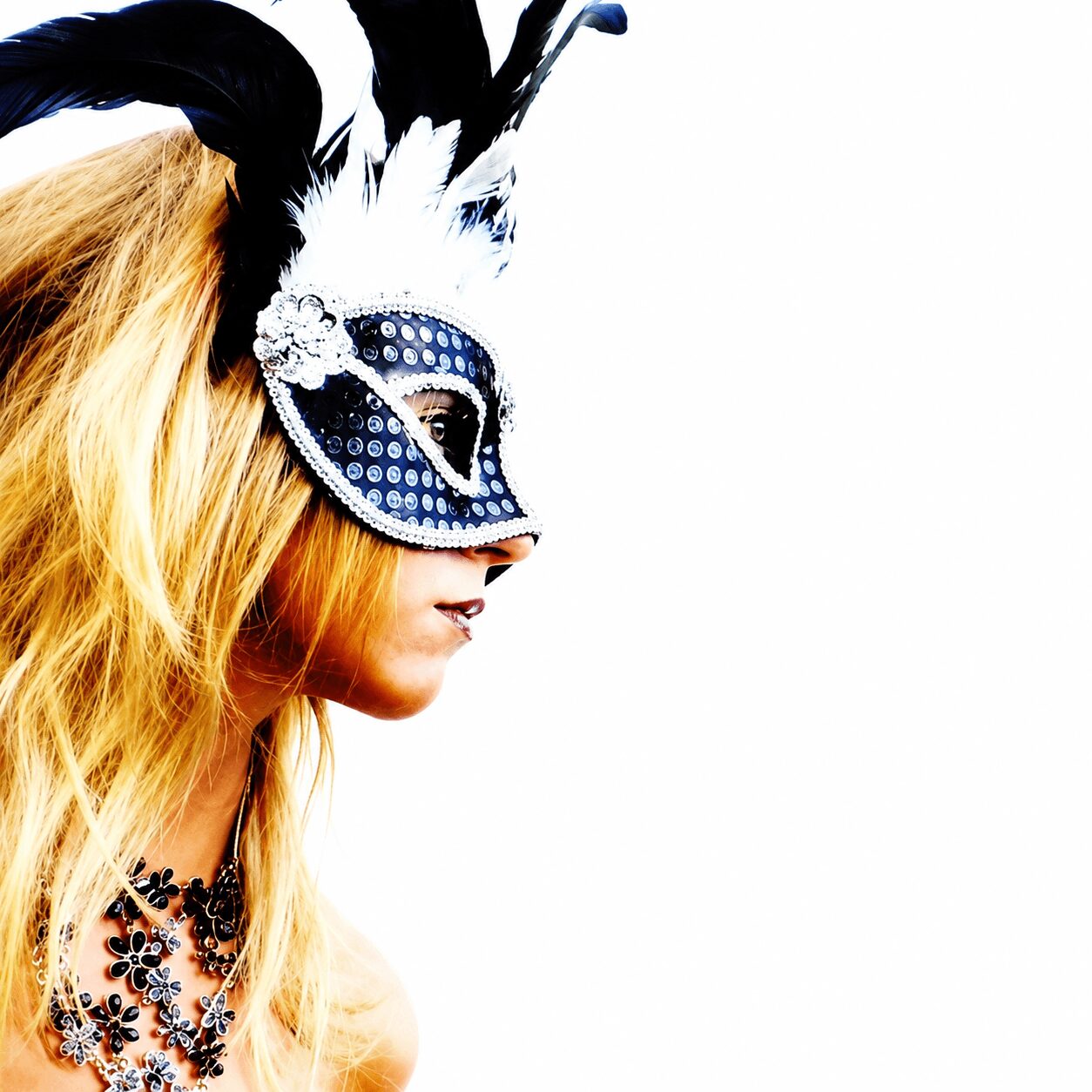 Person wearing a decorative mask with feathers and jewelry, facing left, with long blonde hair, against a white background.