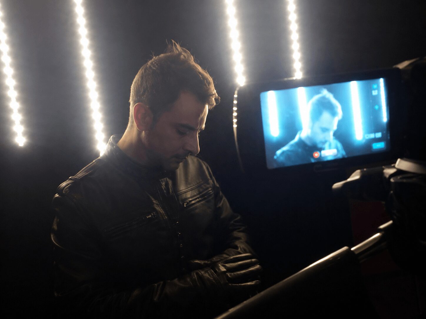 Man with dark hair and beard wearing a black leather jacket, sitting in front of a camera with bright vertical lights behind him.