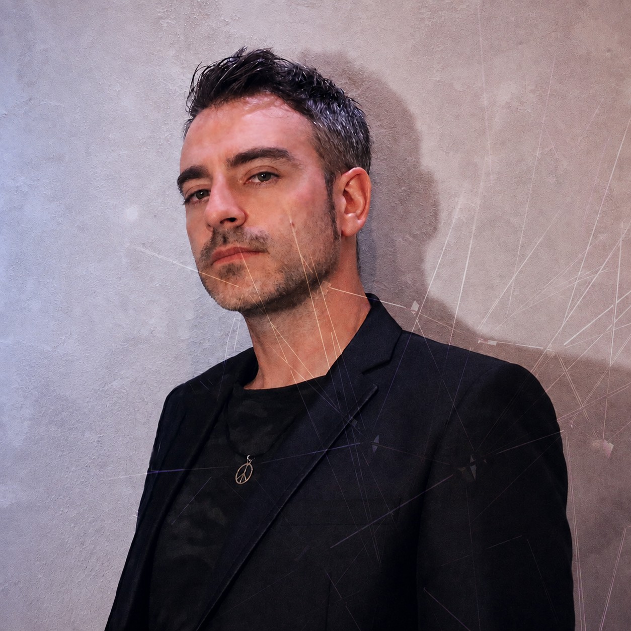 Man with short dark hair and stubble, wearing a black blazer and necklace, standing against a plain wall.