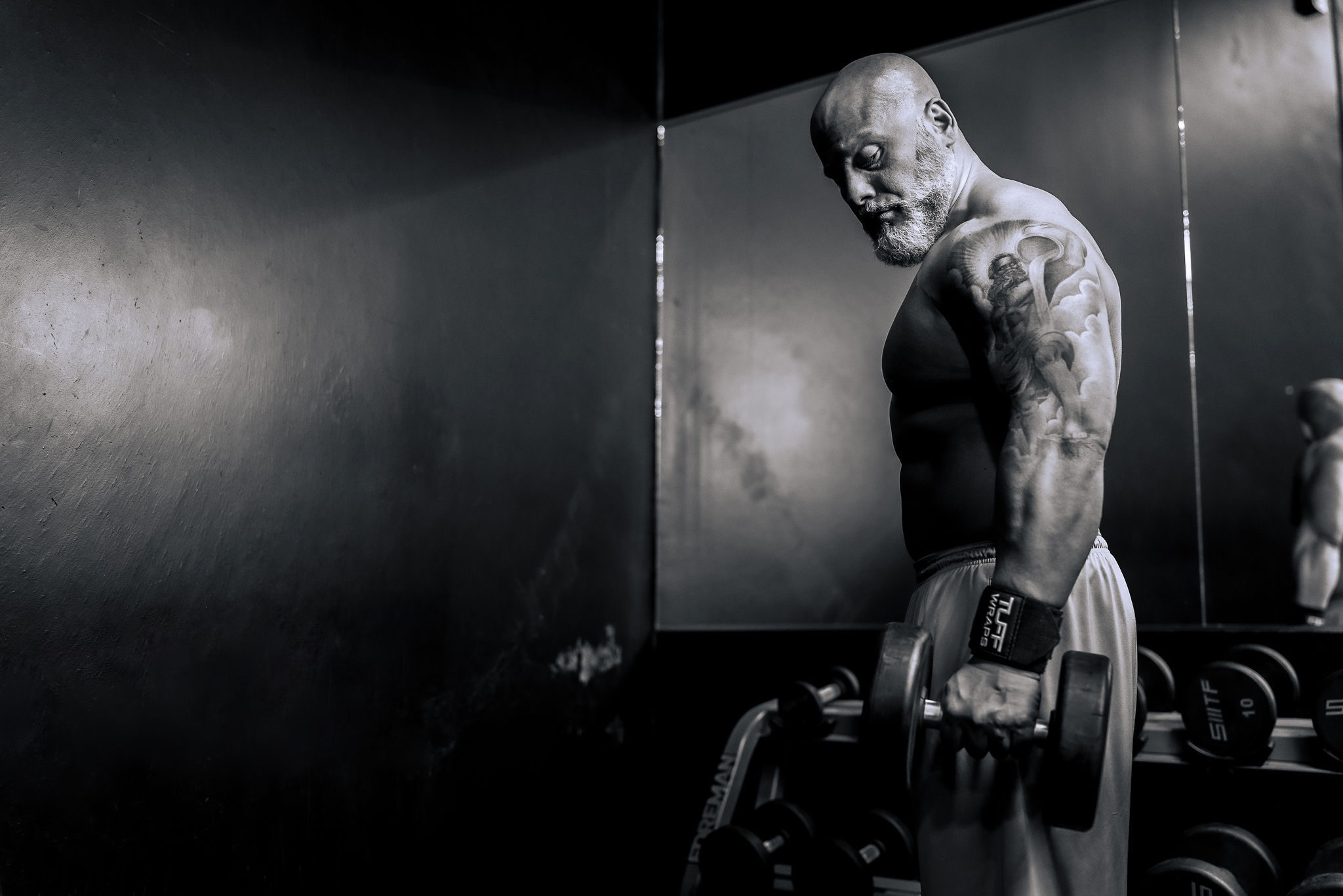 A muscular man with tattoos lifting a dumbbell in a gym, with a mirror reflecting his image.