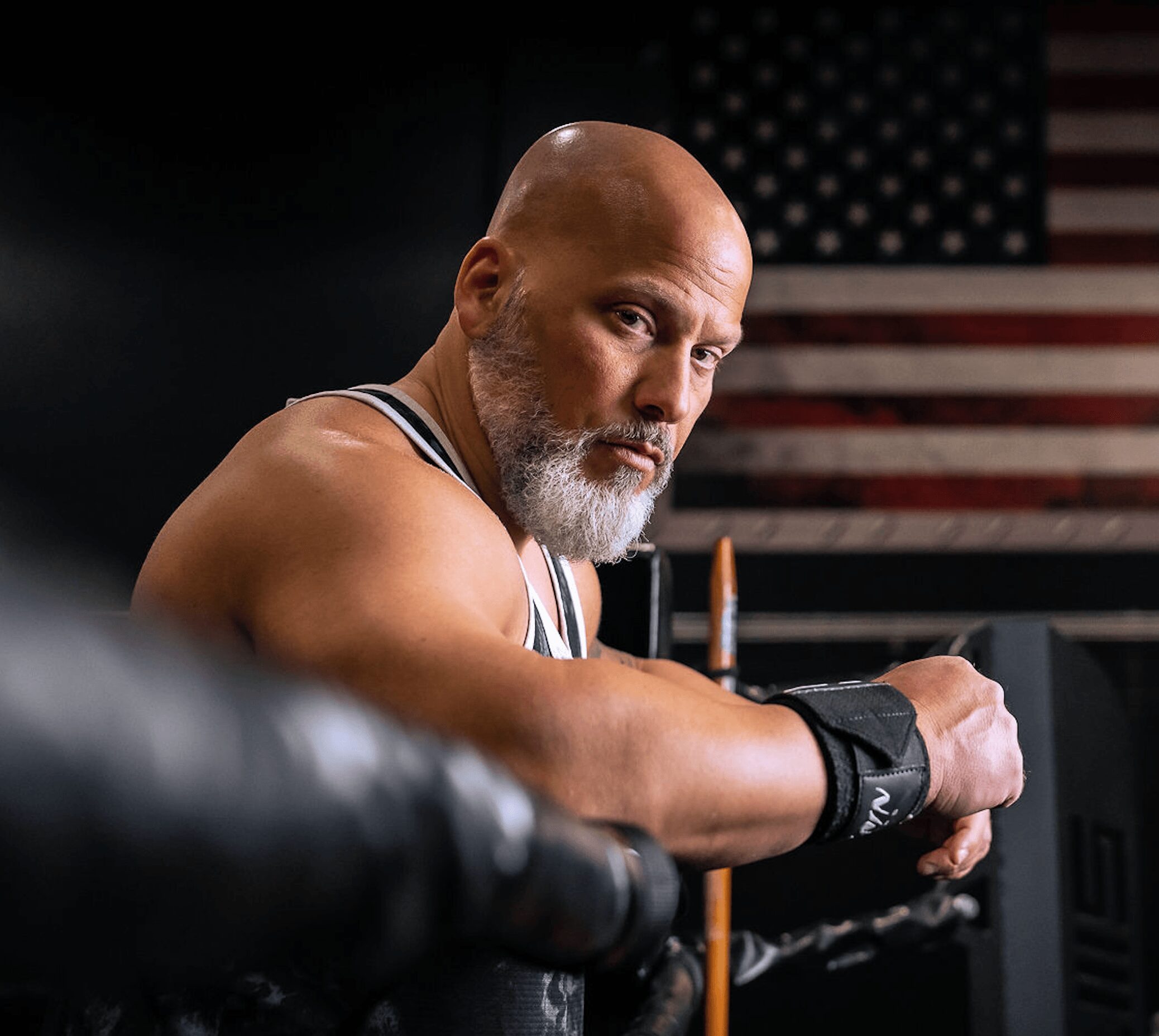 A muscular man with a beard leans on a barbell in a gym, with an American flag in the background.