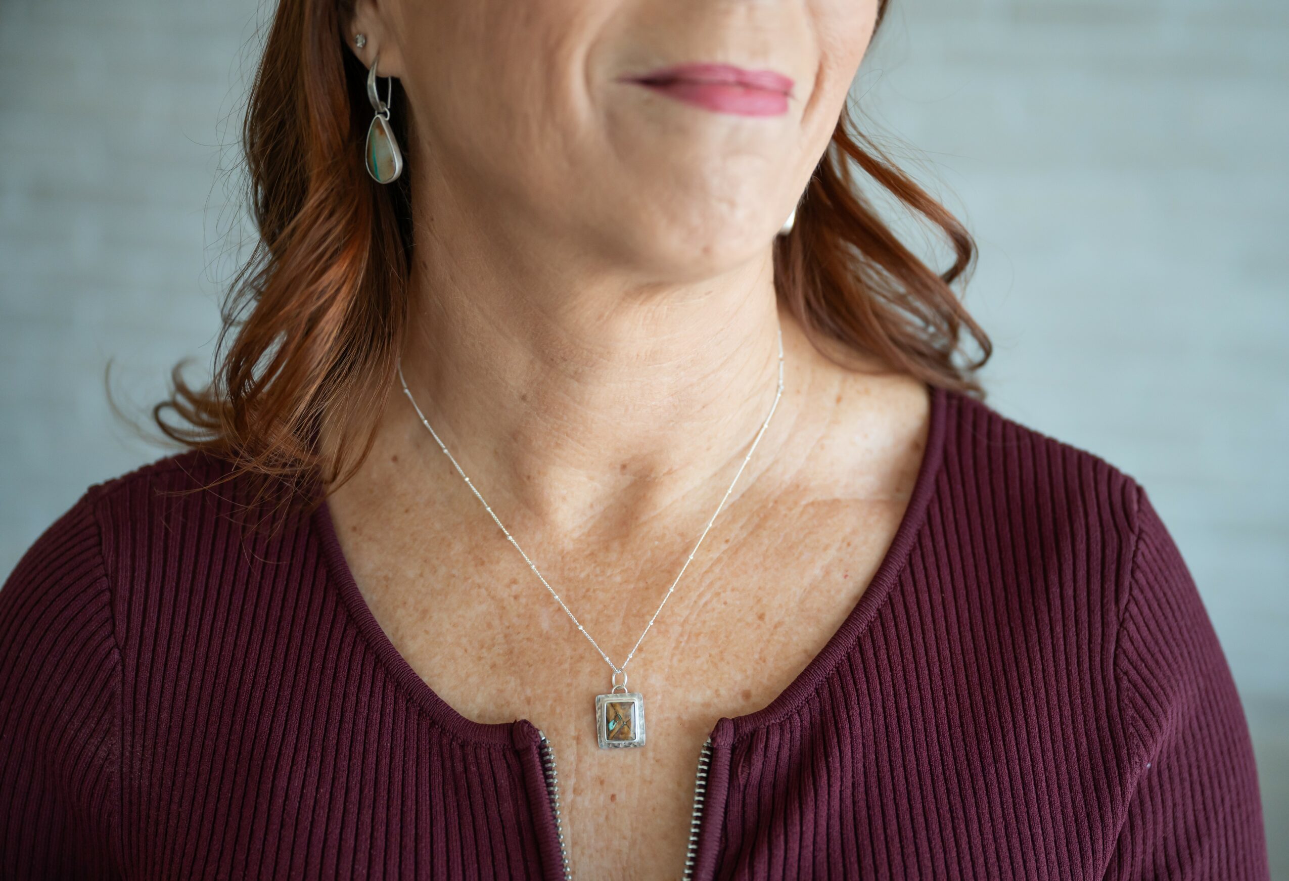 Close-up of a woman wearing a maroon top, earrings, and a necklace with a rectangular pendant, smiling slightly.