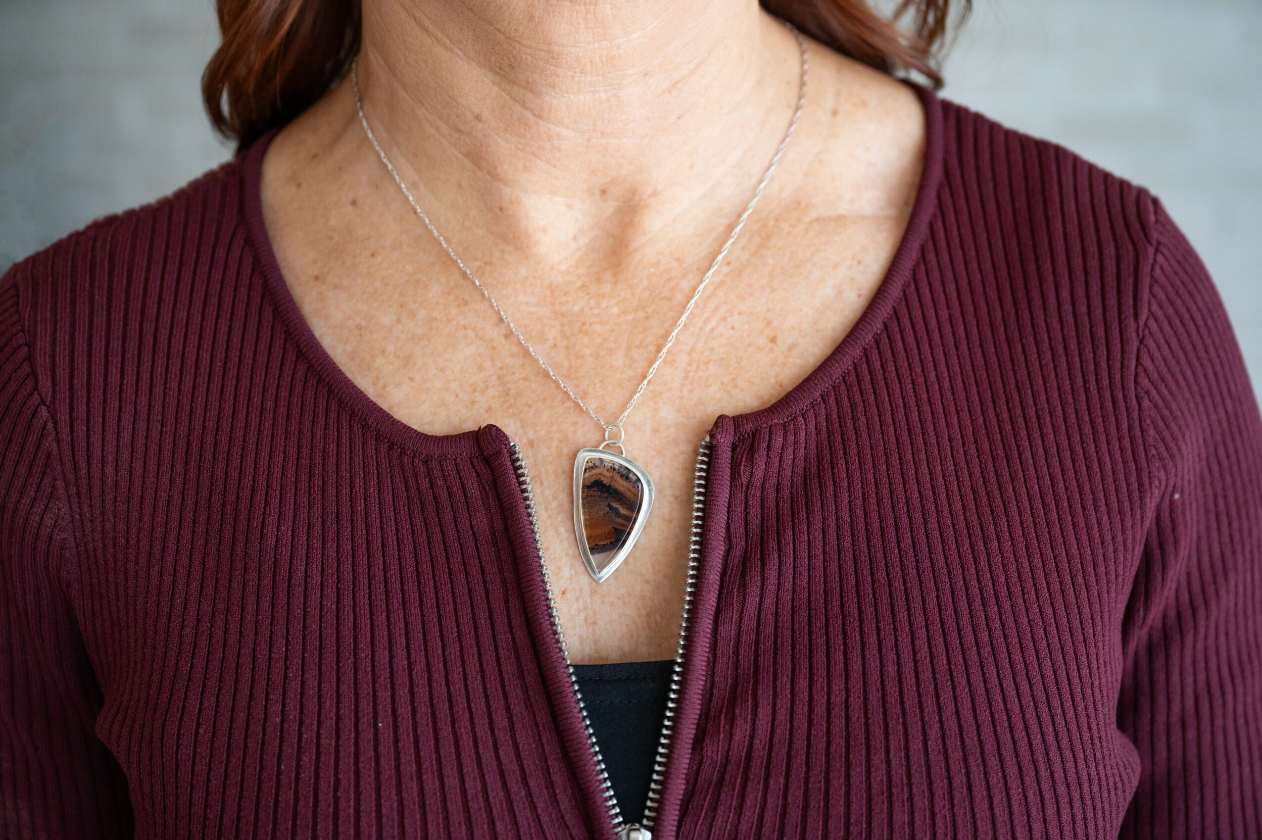 Close-up of a woman wearing a maroon ribbed top with a zipper, necklace with a pendant, and a delicate chain.