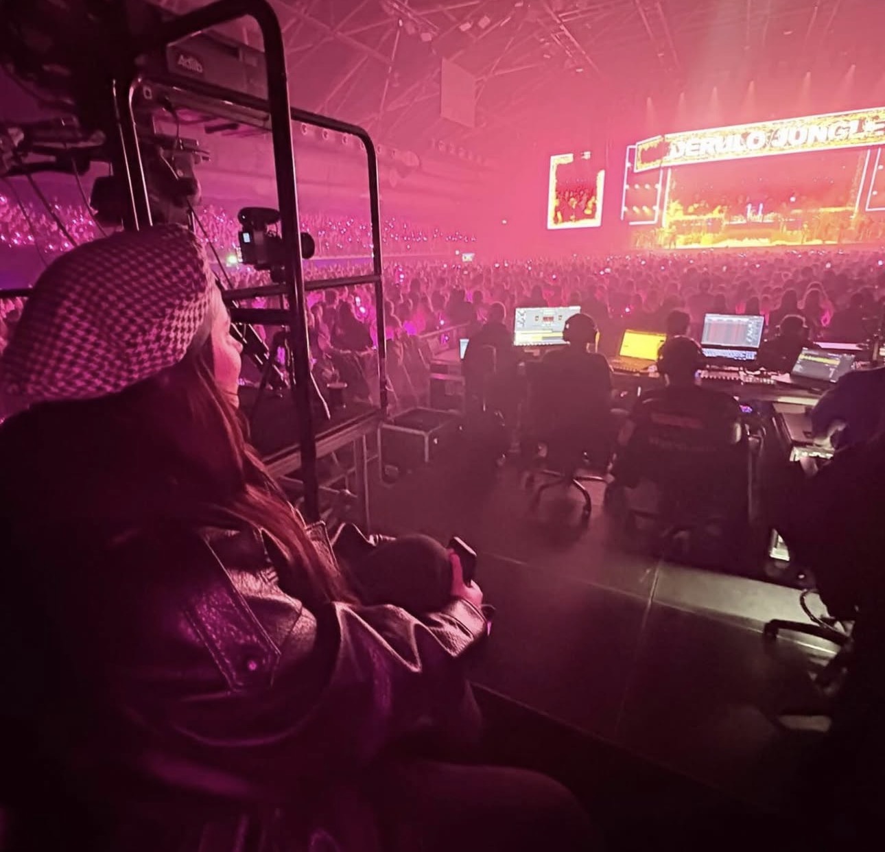 Person sitting in a dark room with a large screen and equipment, illuminated by pink and purple lights, at an event or concert.
