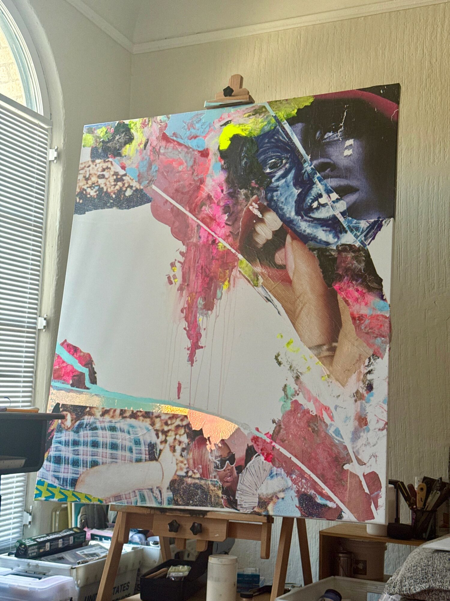 Large colorful abstract painting with a partially visible face, on an easel in a room with a window and various objects.
