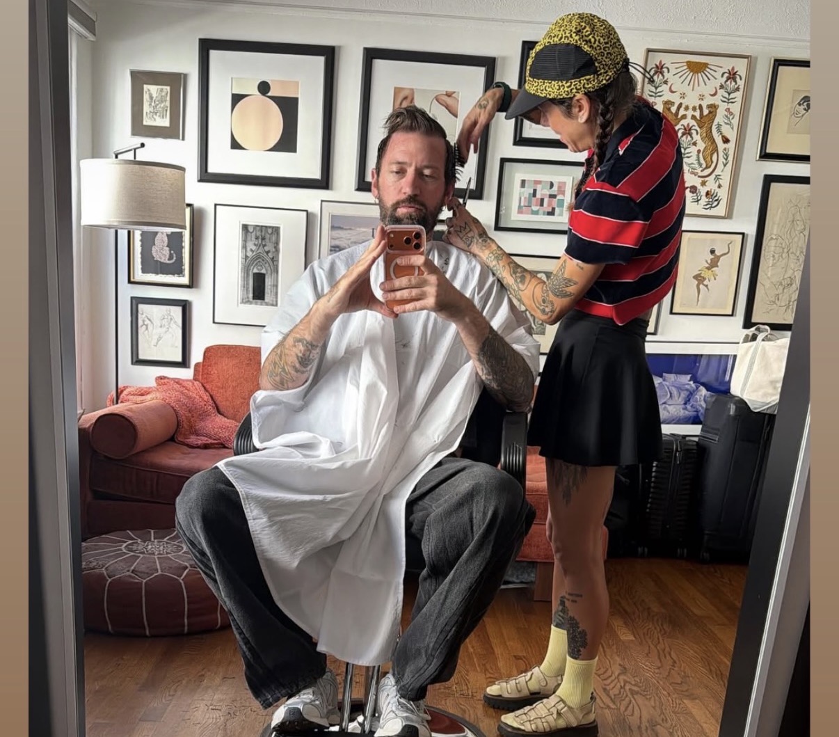 Man in a barber cape sitting in a chair, woman styling his hair, art gallery wall in background.