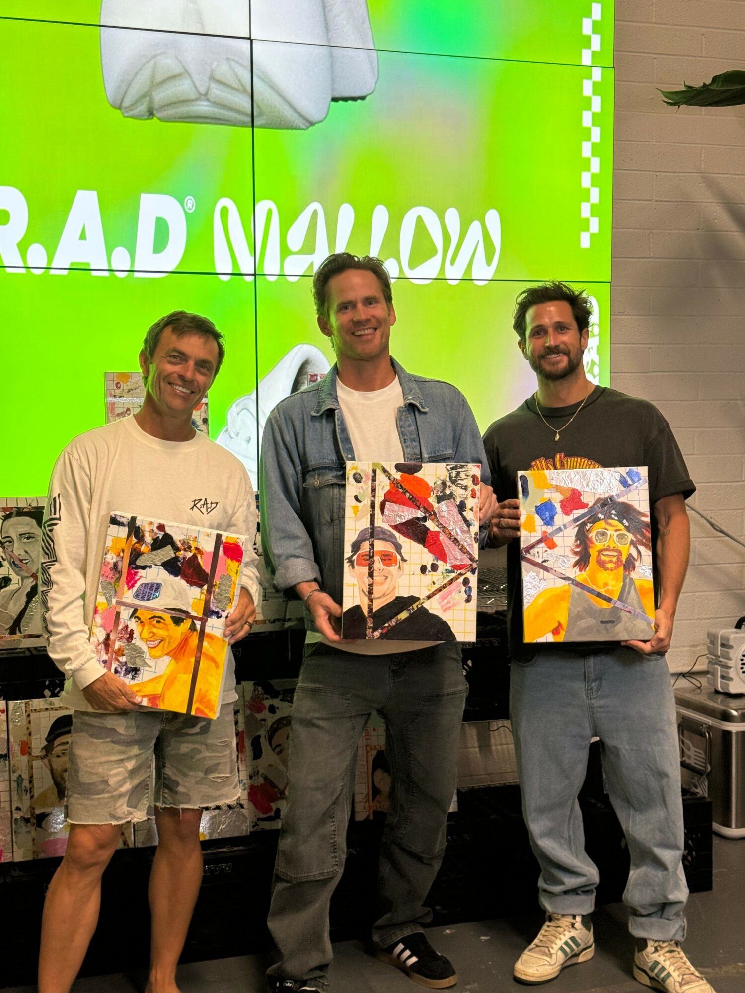 Three people holding colorful paintings in front of a green wall with white text, standing indoors. One person is smiling, others are looking at the camera.