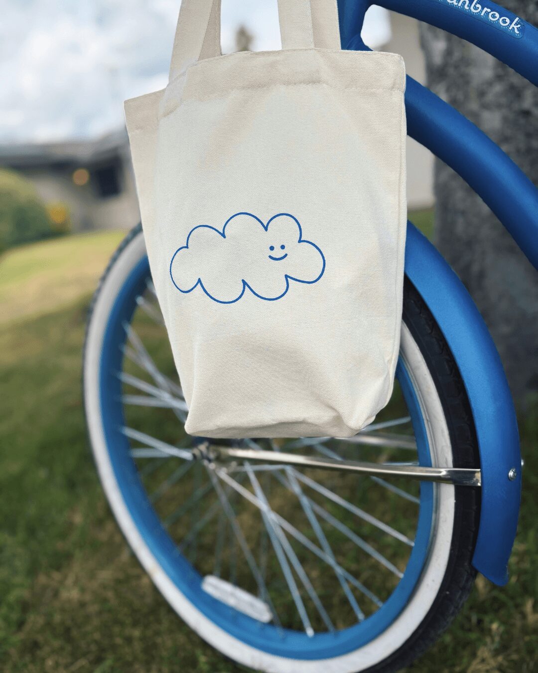 Canvas bag with a smiling cloud drawing hanging on a blue bicycle wheel outdoors.