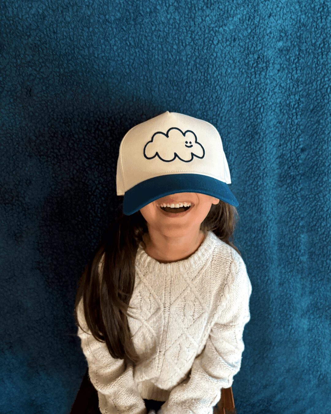 Child wearing a white cap with a cloud drawing, smiling, against a blue textured background.