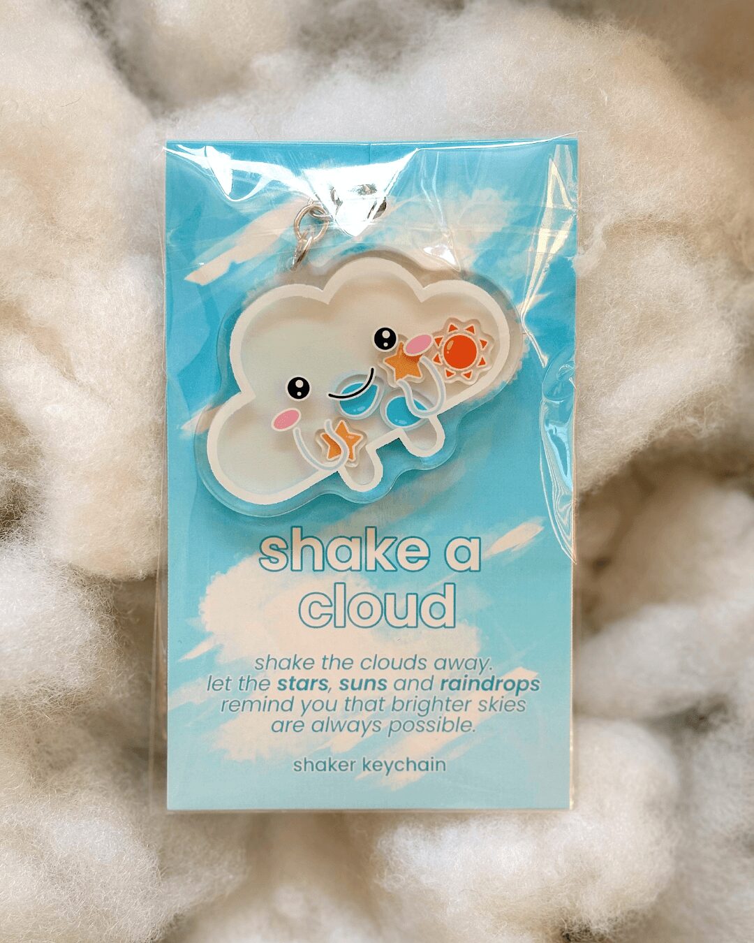 Cloud-shaped keychain with smiling face, pink cheeks, and a small rainbow, in blue packaging with white text.