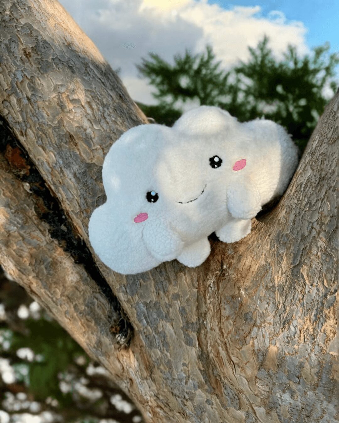 A cute plush cloud with a smiling face, pink cheeks, and small arms, resting on a tree branch outdoors.