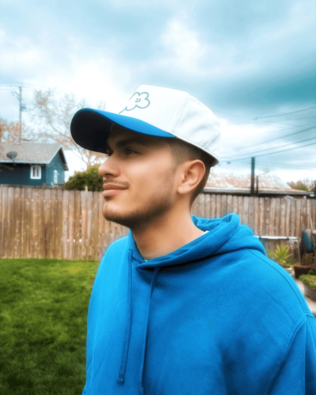 Young man in a blue hoodie and white cap outdoors, with a wooden fence and cloudy sky in background.