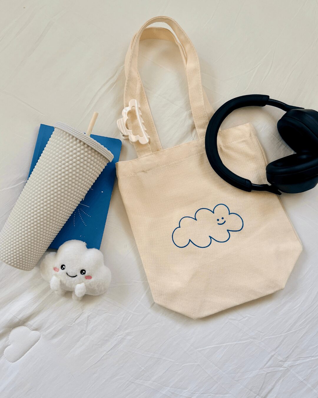 Cream-colored tote bag with a cloud drawing, black headphones, a small plush cloud, a blue water bottle, and a gray mesh item on a white surface.