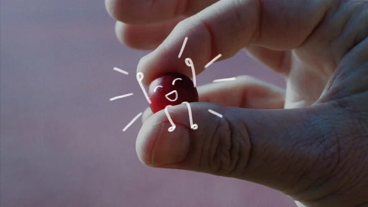 Hand holding a cherry with a smiling face and doodle-like lines, against a blurred background.