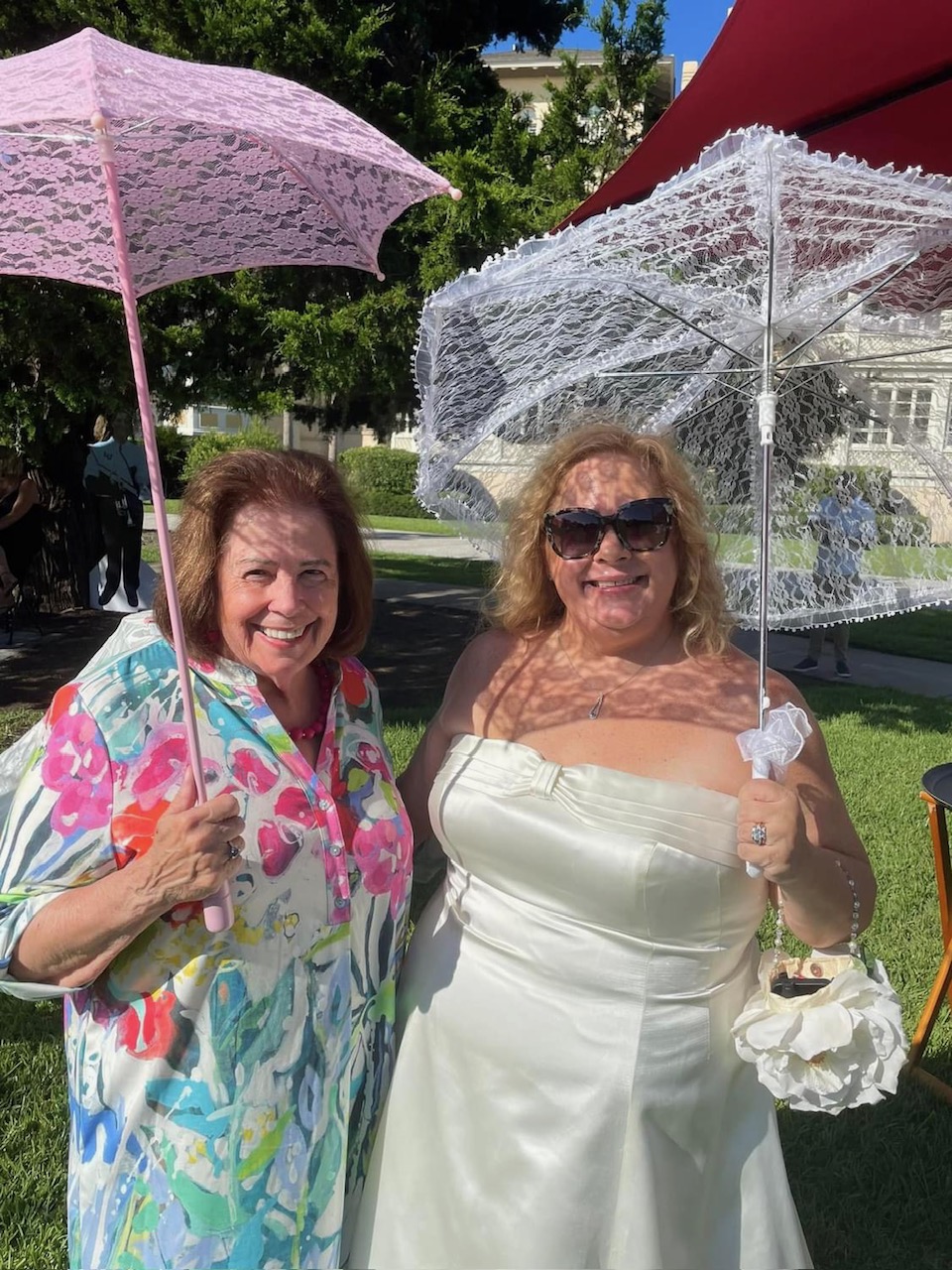 Two women holding umbrellas outdoors on sunny day, smiling, with trees and buildings in background.