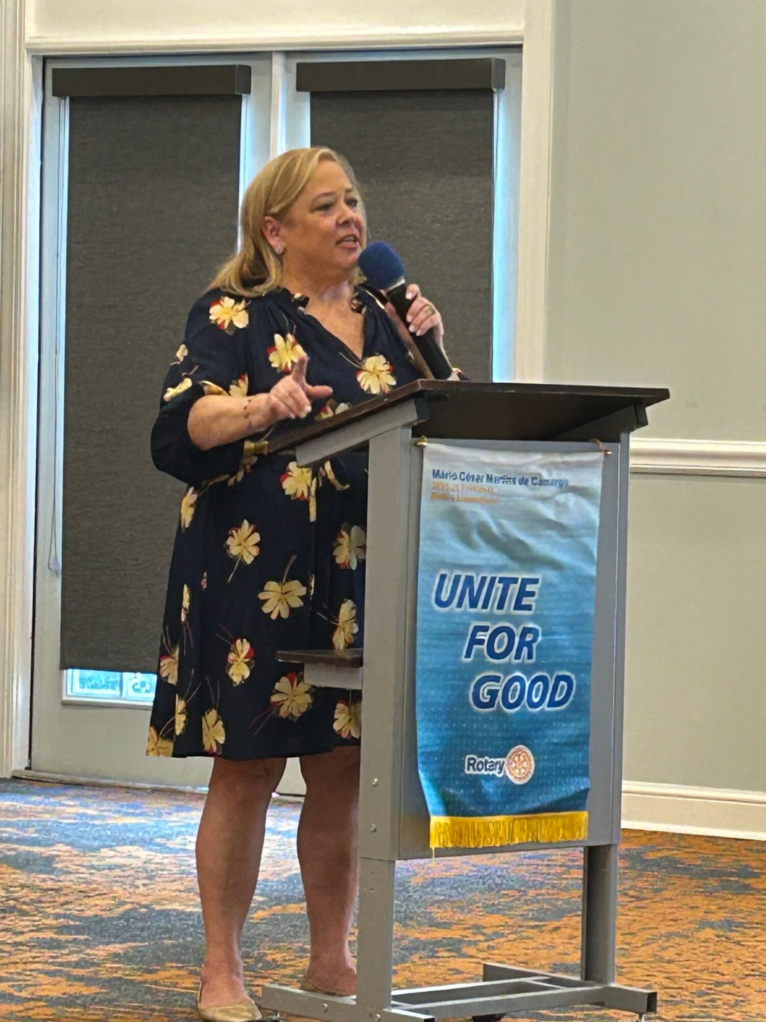 Woman speaking at a podium with a banner that reads 'Unite for Good', in a room with windows and carpeted floor.