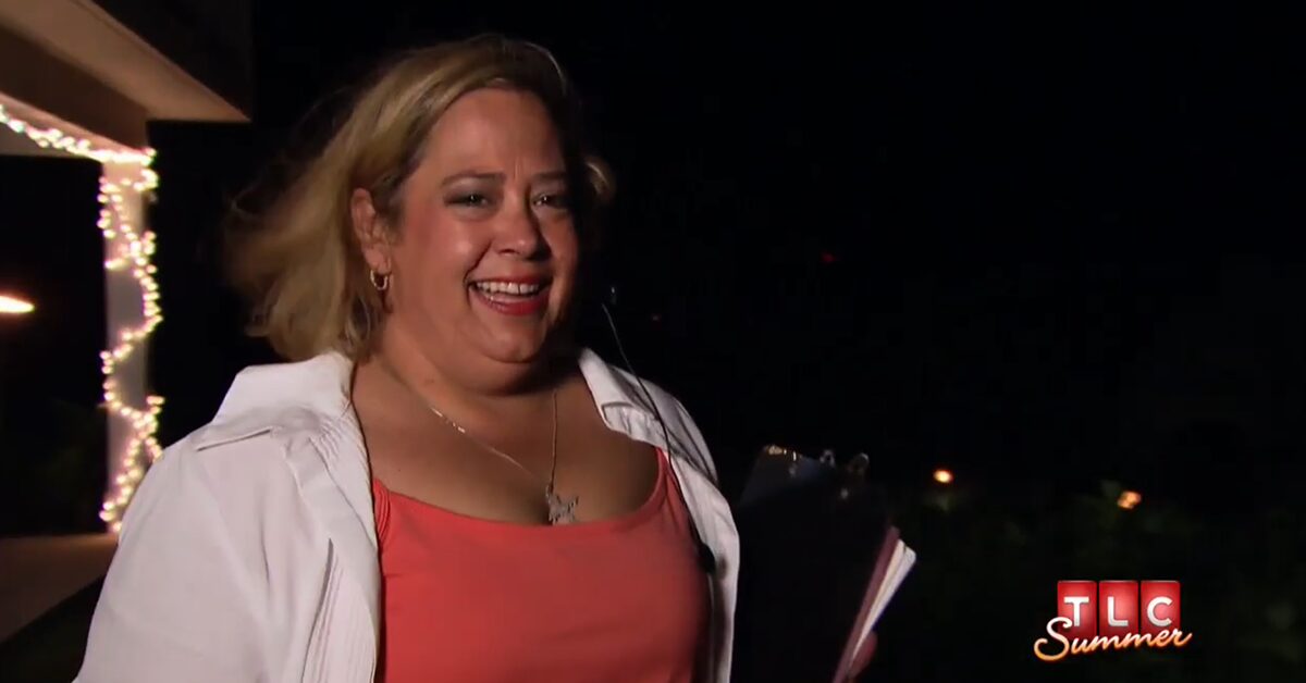 Woman smiling outdoors at night, wearing a white jacket and coral top, holding a notebook, with lights in the background.