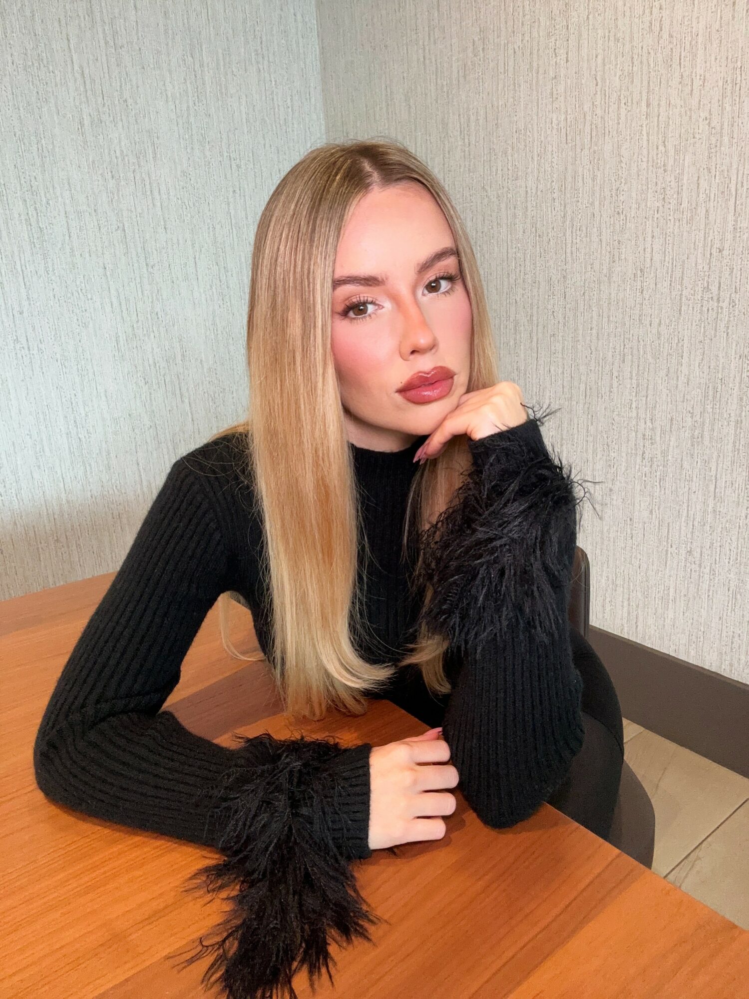 Young woman with long blonde hair, wearing a black sweater with furry cuffs, sitting at a wooden table, resting her chin on her hand.