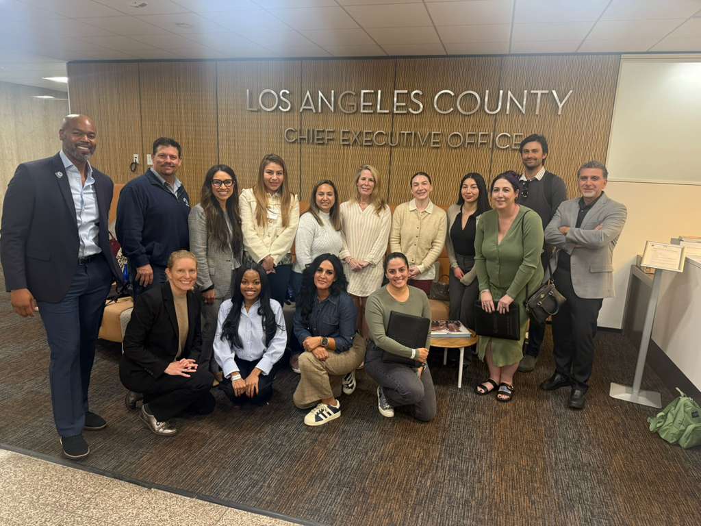 Group of 14 people standing and sitting in front of Los Angeles County Chief Executive Office sign, smiling.