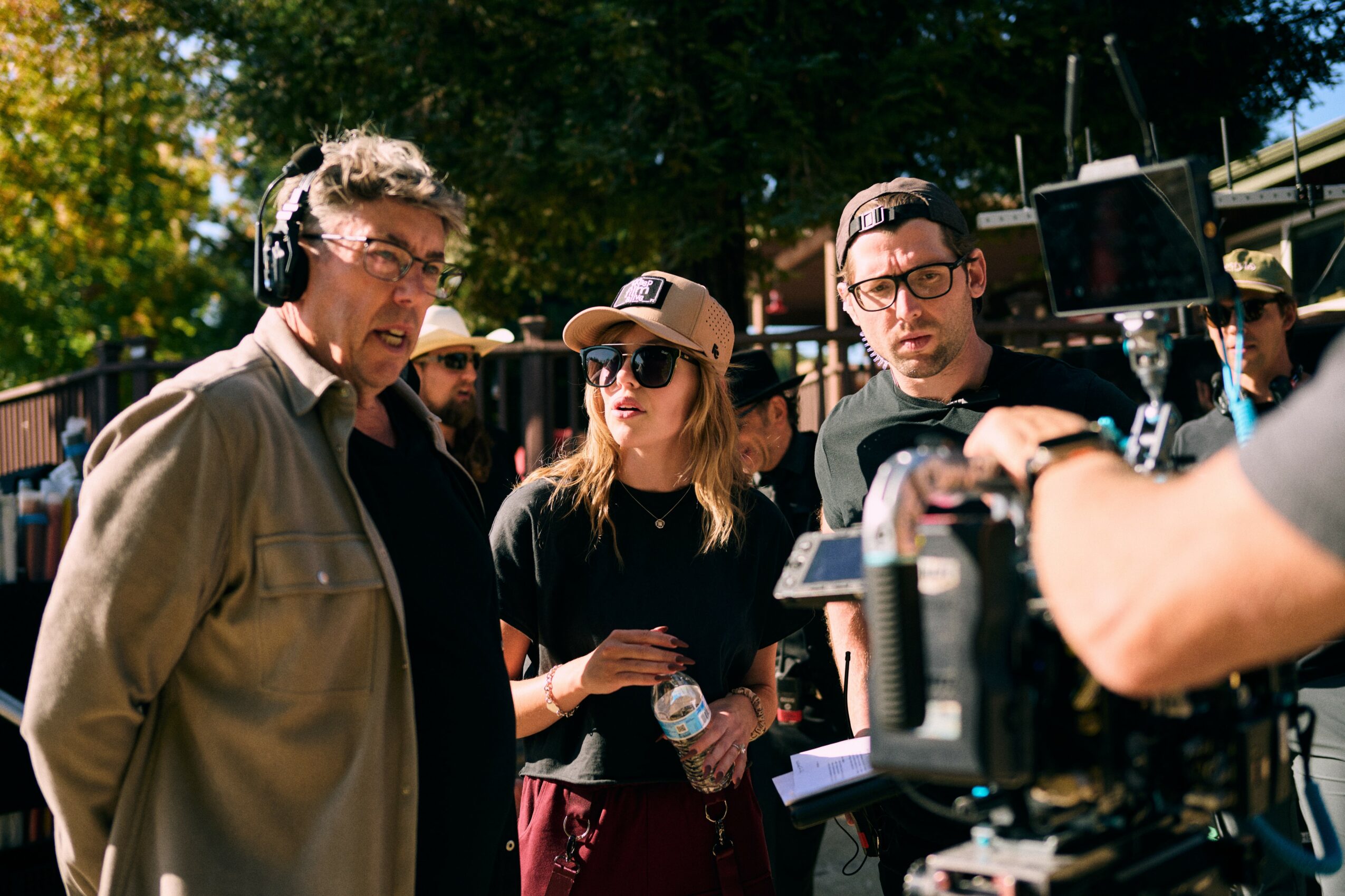 Three people outdoors, one with headphones, another with sunglasses and a cap, and a third with glasses, near camera equipment.