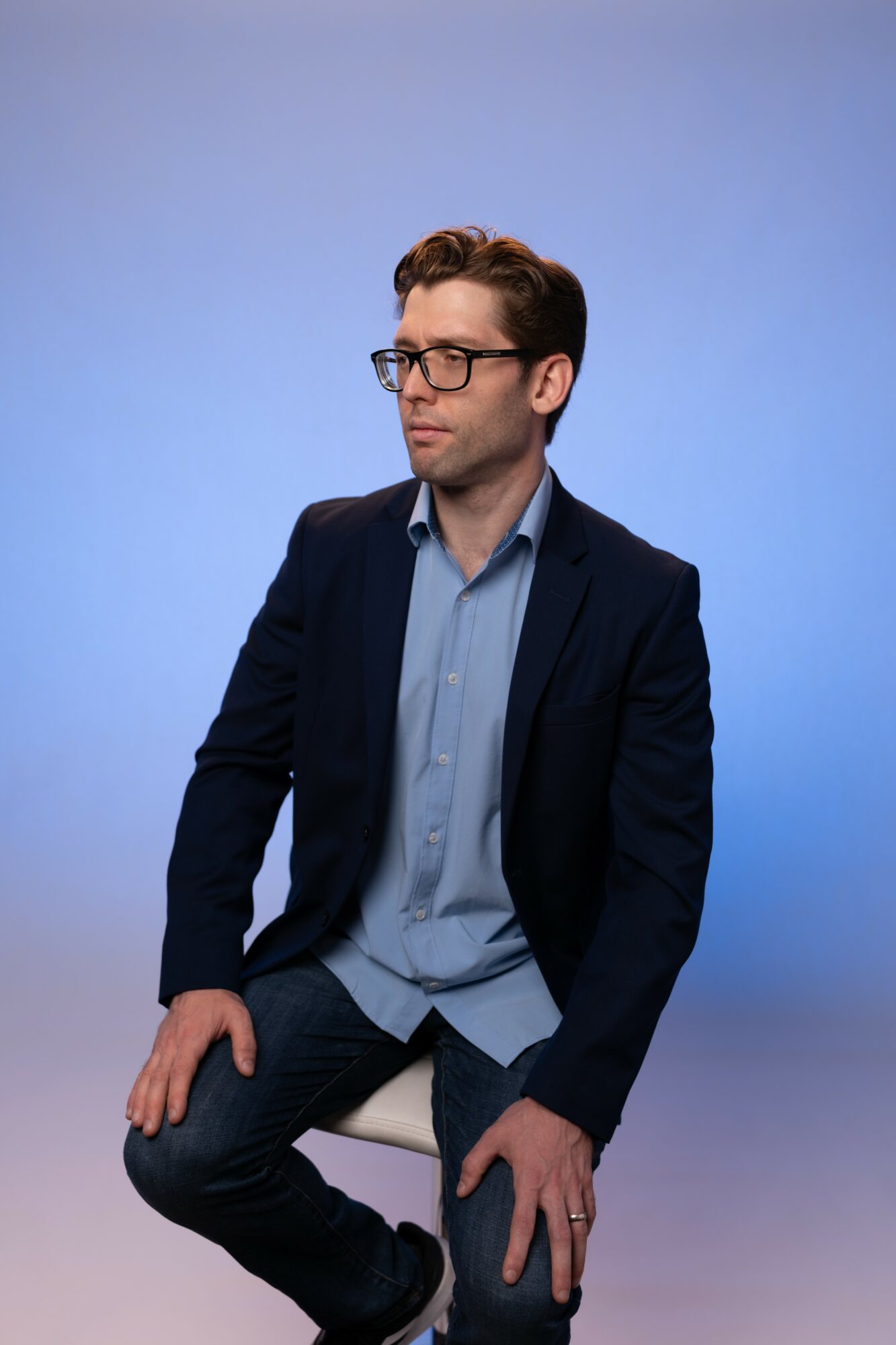 Man with glasses in a dark blazer and light blue shirt, sitting against a blue background.