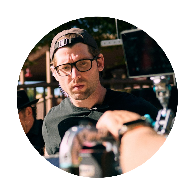 Man with glasses and a cap looks at camera outdoors, with equipment and a person in the foreground.