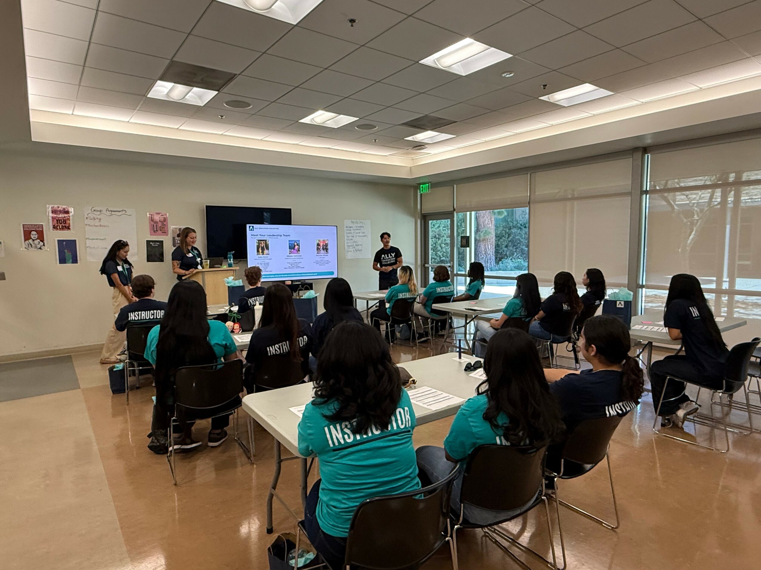 Group of people seated in a classroom watching a presentation on a large screen, with a few standing near the screen.