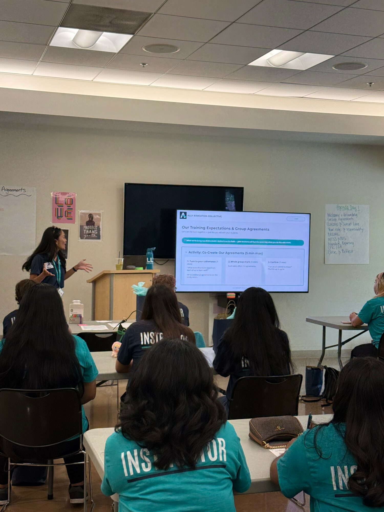 Classroom with students facing a large screen, teacher at front, students wearing teal shirts, some taking notes.