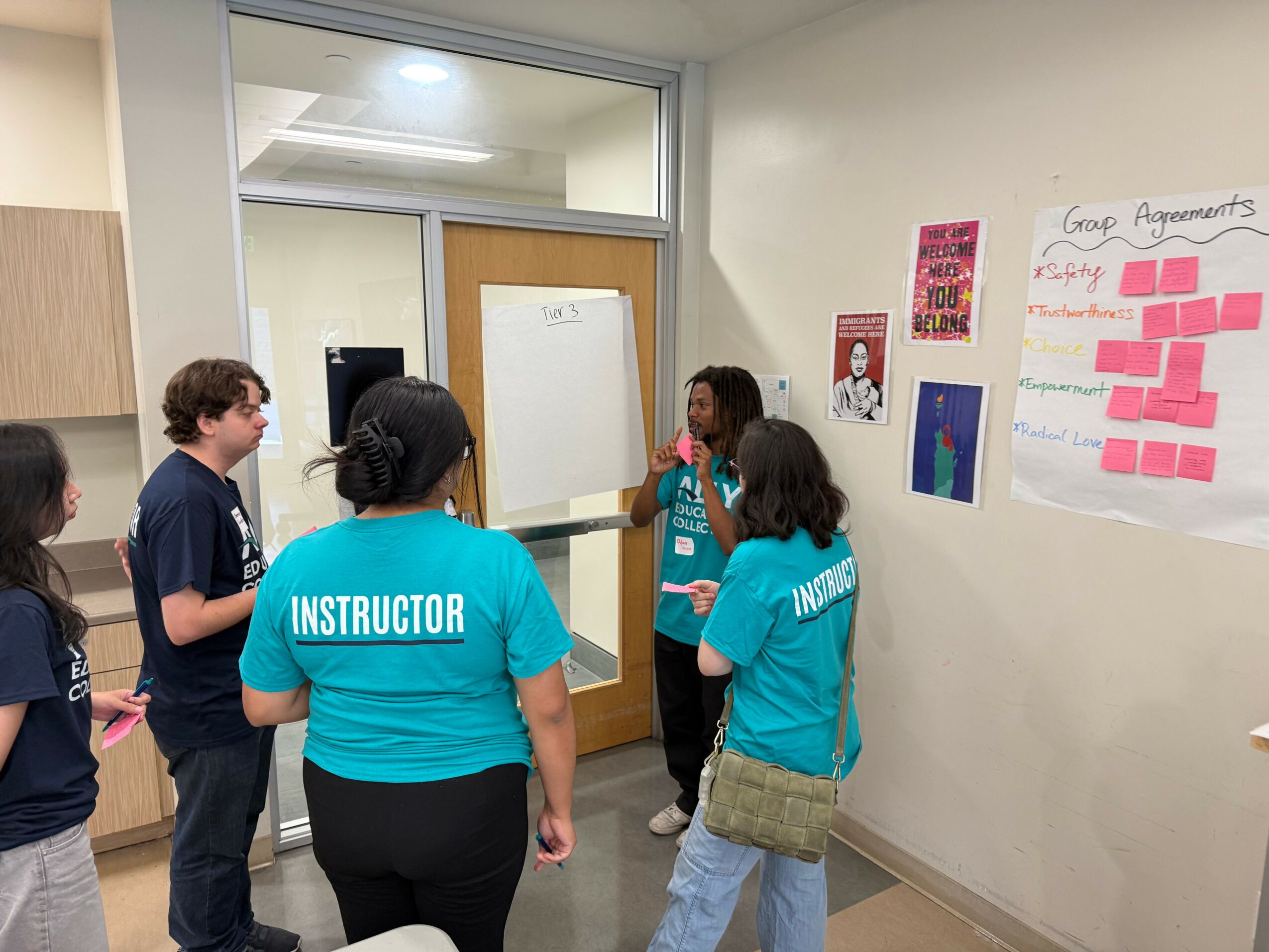 Group of people, including instructor, gathered in a classroom around a whiteboard, with posters on the wall and a door in the background.