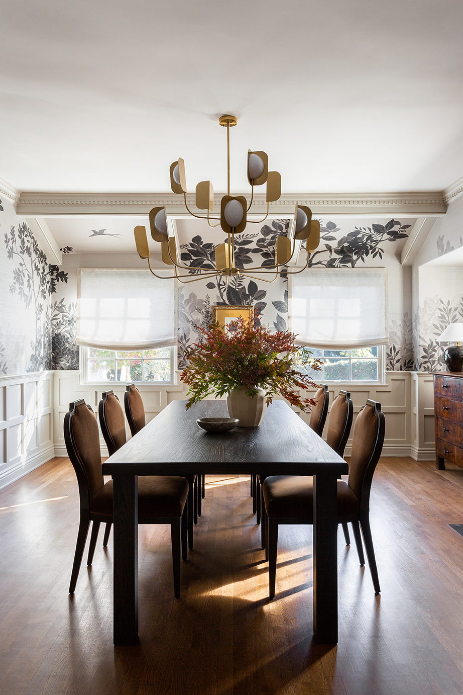 Dining room with a black table, eight chairs, a floral centerpiece, chandelier, and large windows with curtains.