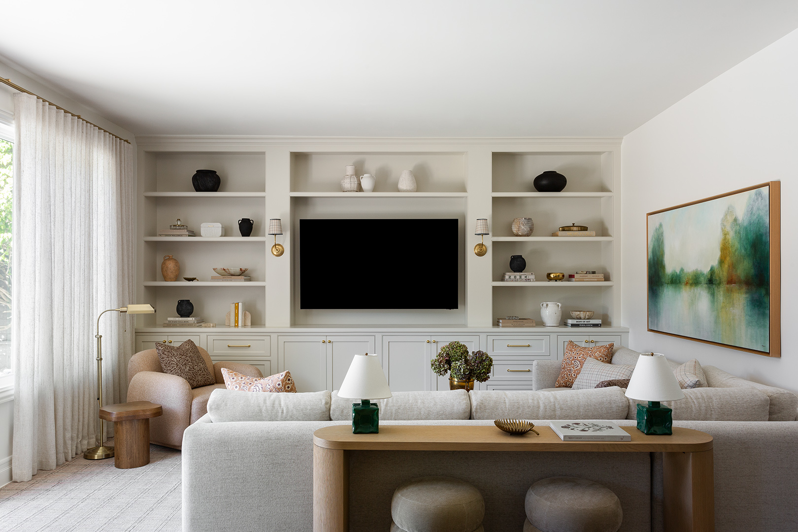 Living room with white shelves, a large TV, two sofas, and a painting on the wall, with natural light from windows.
