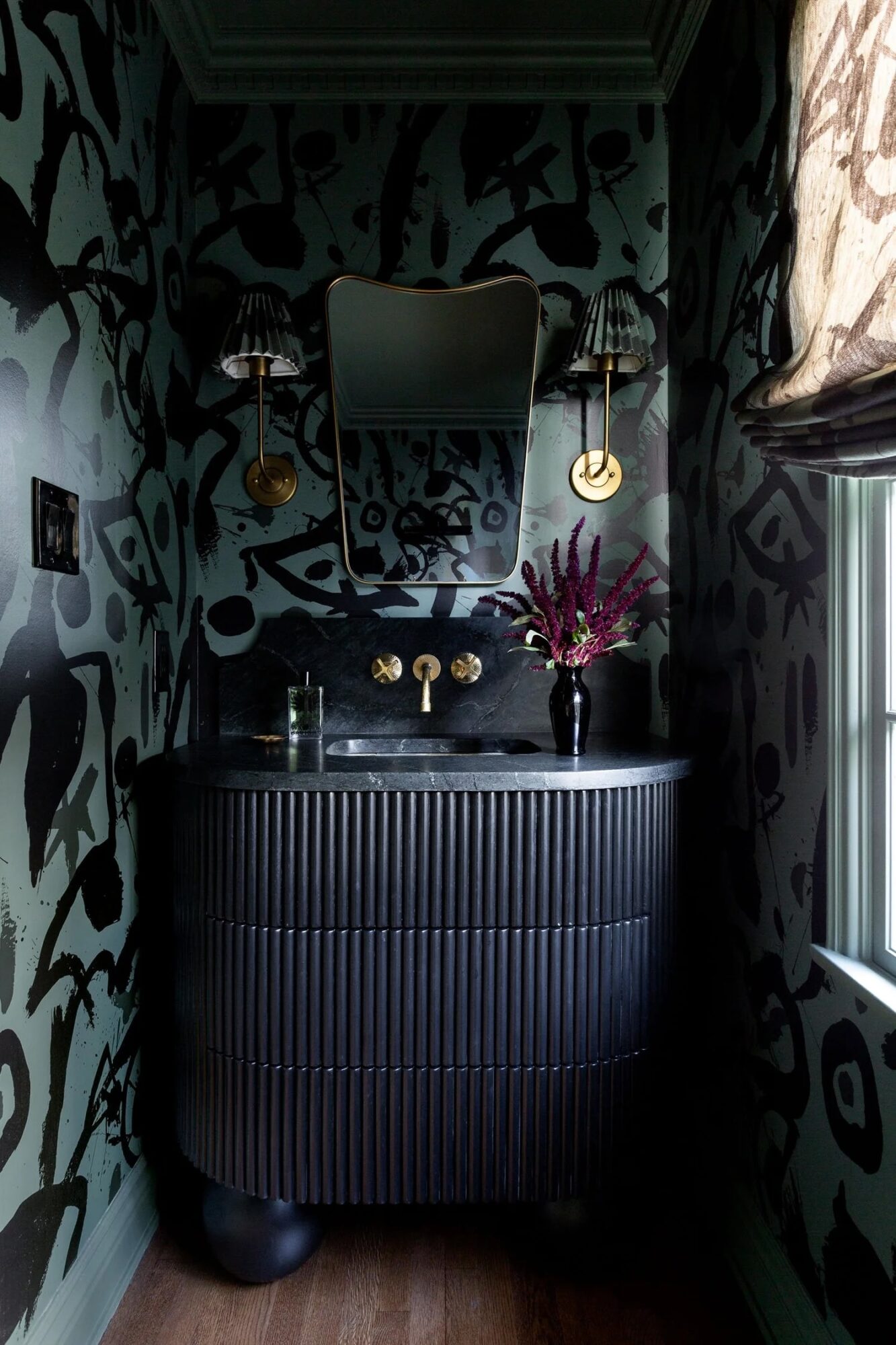 Bathroom with dark patterned wallpaper, black sink with gold fixtures, mirror, and a vase with purple flowers, window on right.