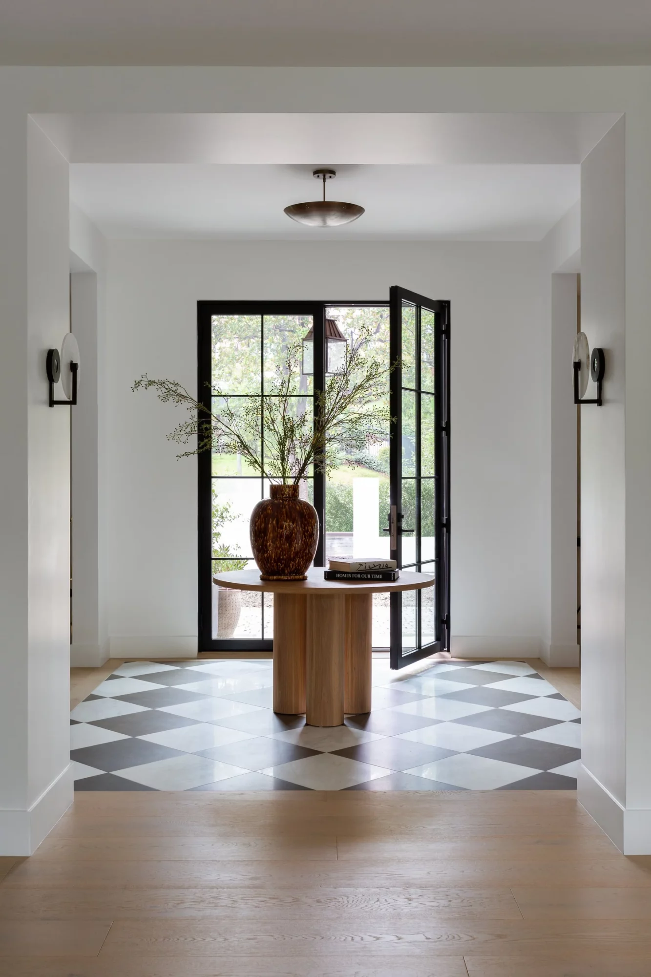 View through a doorway to a room with a round table, a large vase with branches, and open glass doors showing trees outside.