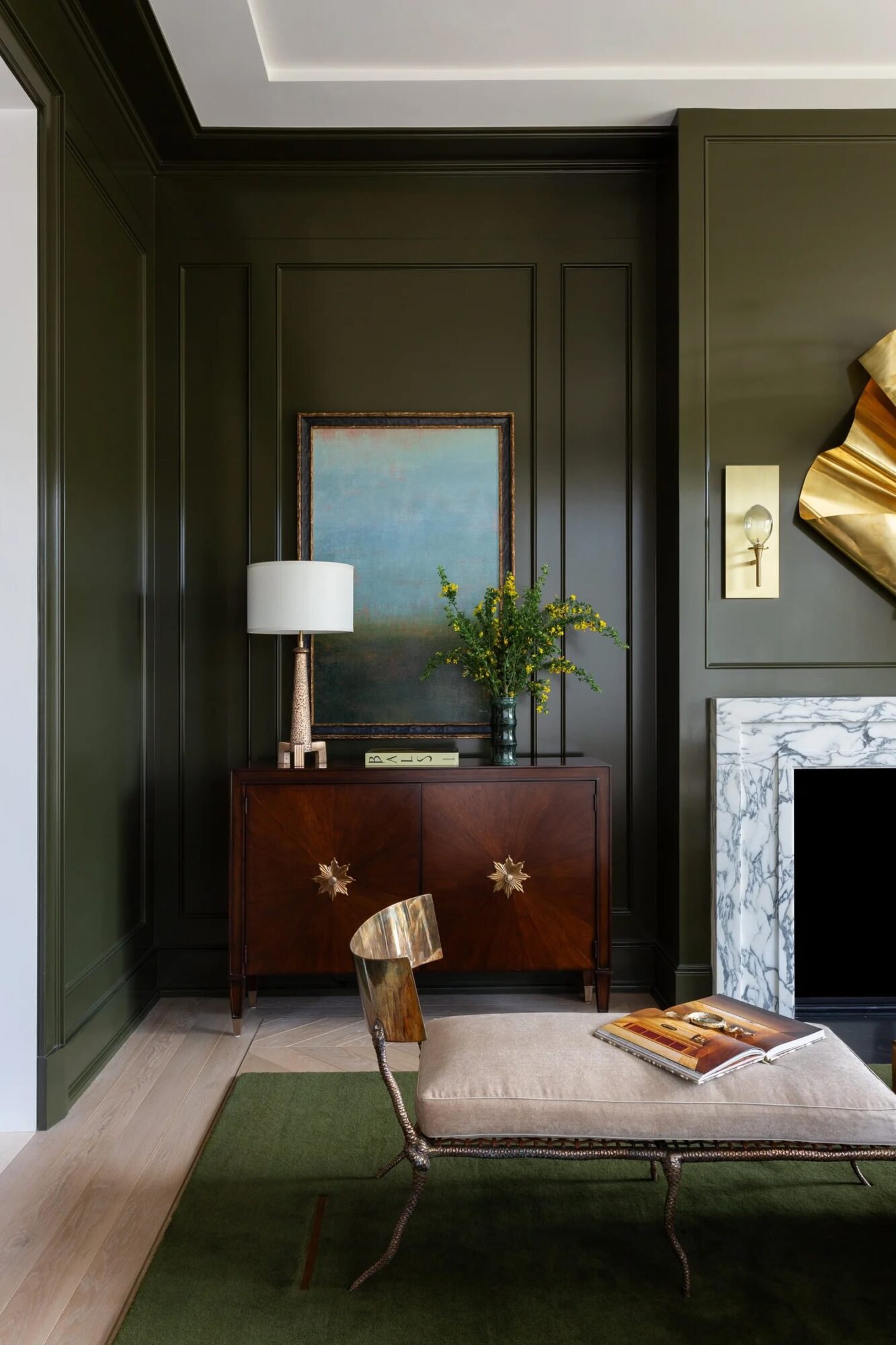 Interior corner with dark green walls, a wooden sideboard, a table lamp, a framed painting, and a plant, with a marble fireplace and a chair with a book.