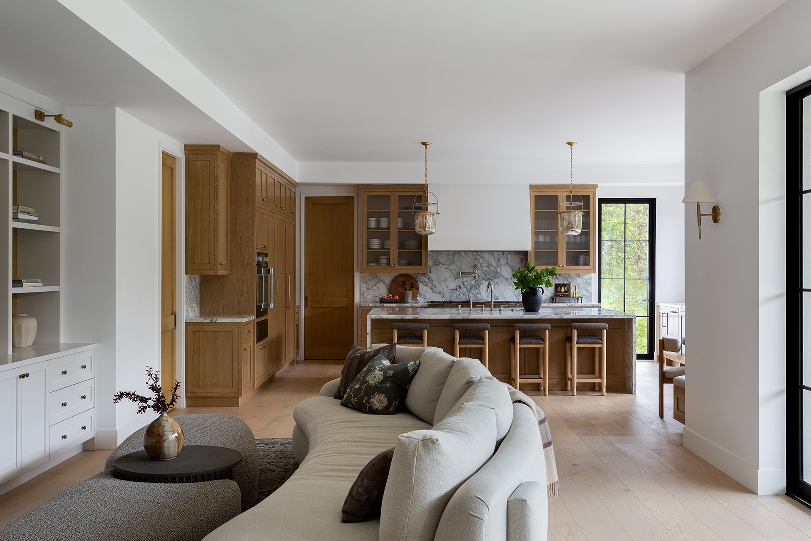 Living room with white walls, wooden kitchen cabinets, a kitchen island, and a white sofa with pillows, sunlight through windows.