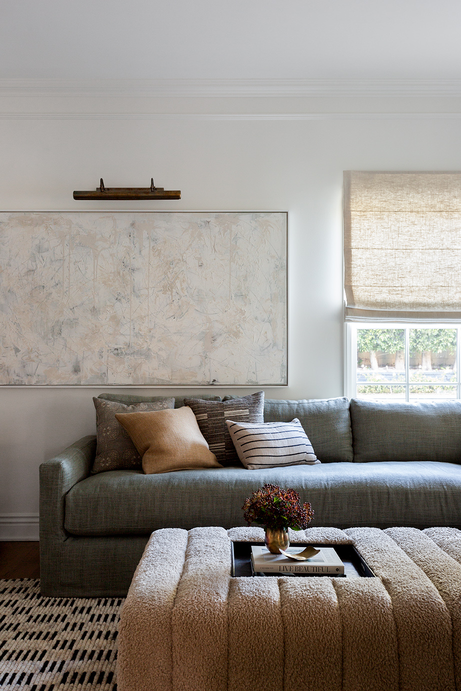 Living room with a gray sofa, beige ottoman, window with a shade, and a wall with artwork and a small shelf.
