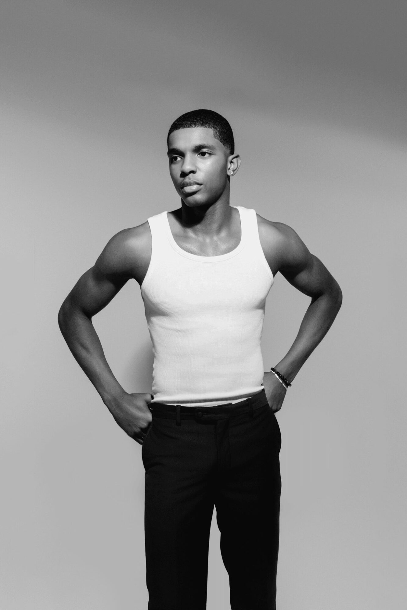 Young man in tank top and pants standing outdoors with hands on hips, looking to the side, clear sky background.