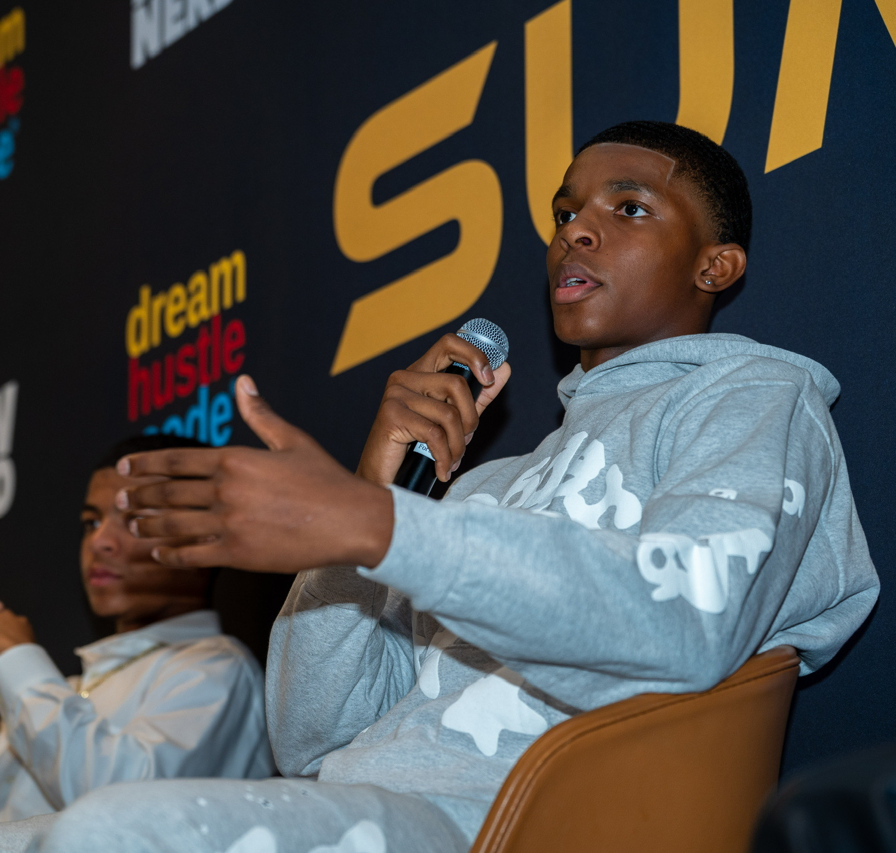 Young person speaking into a microphone, wearing a gray hoodie, seated on a brown chair, with a dark background and text.