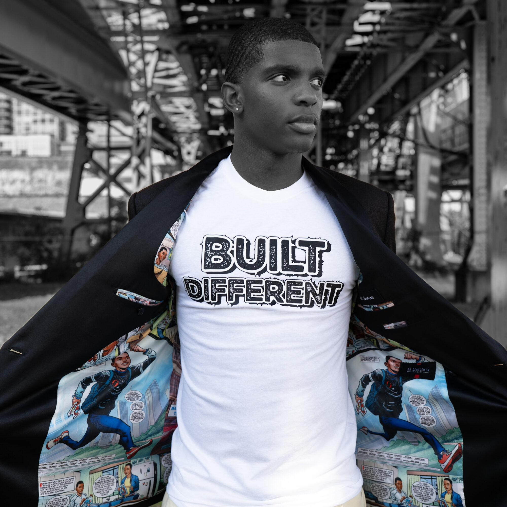 Young man standing outdoors under a bridge, wearing a white T-shirt with text and an open jacket, looking to the side.