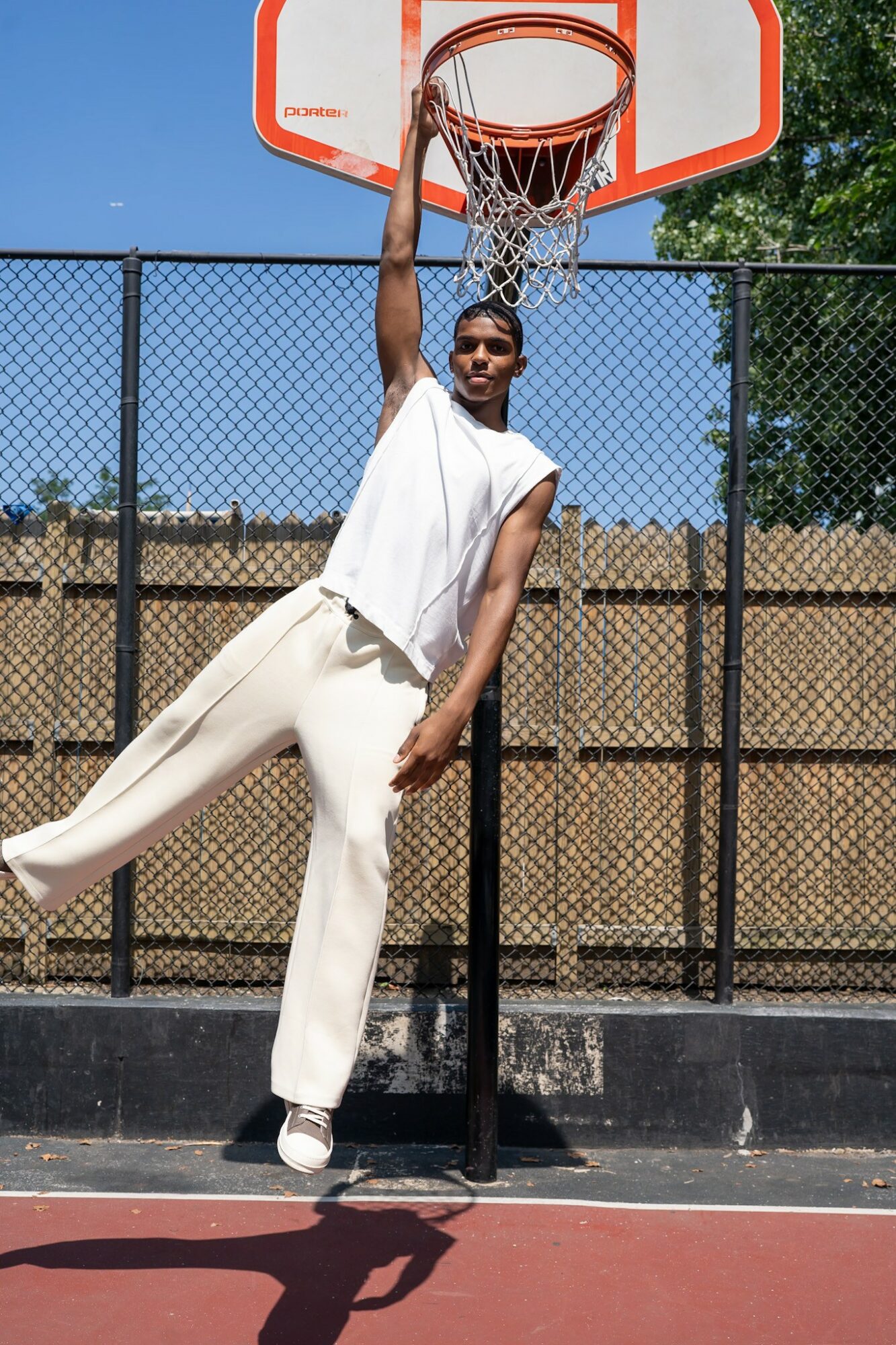 Person hanging from basketball hoop with one hand, on outdoor court, wearing white outfit, smiling, sunny day.