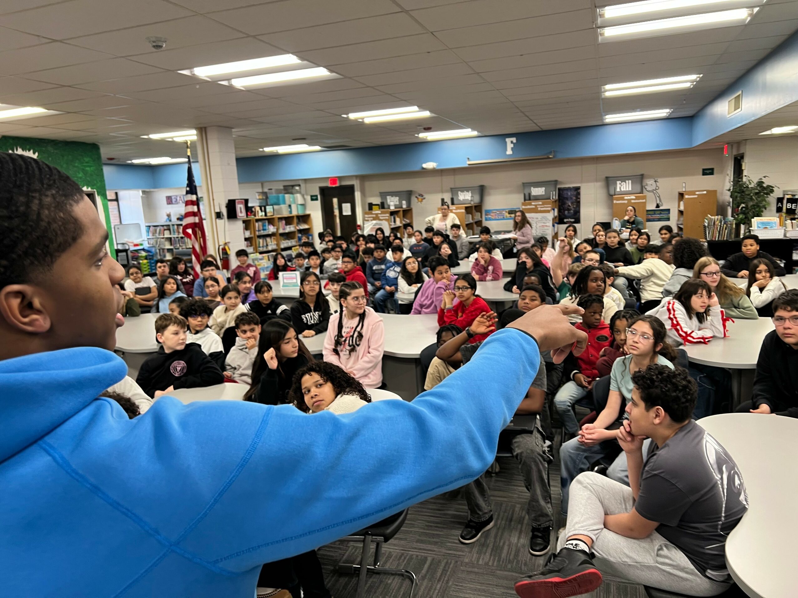 A person in a blue hoodie is speaking to a large group of students seated at tables in a library or classroom setting.