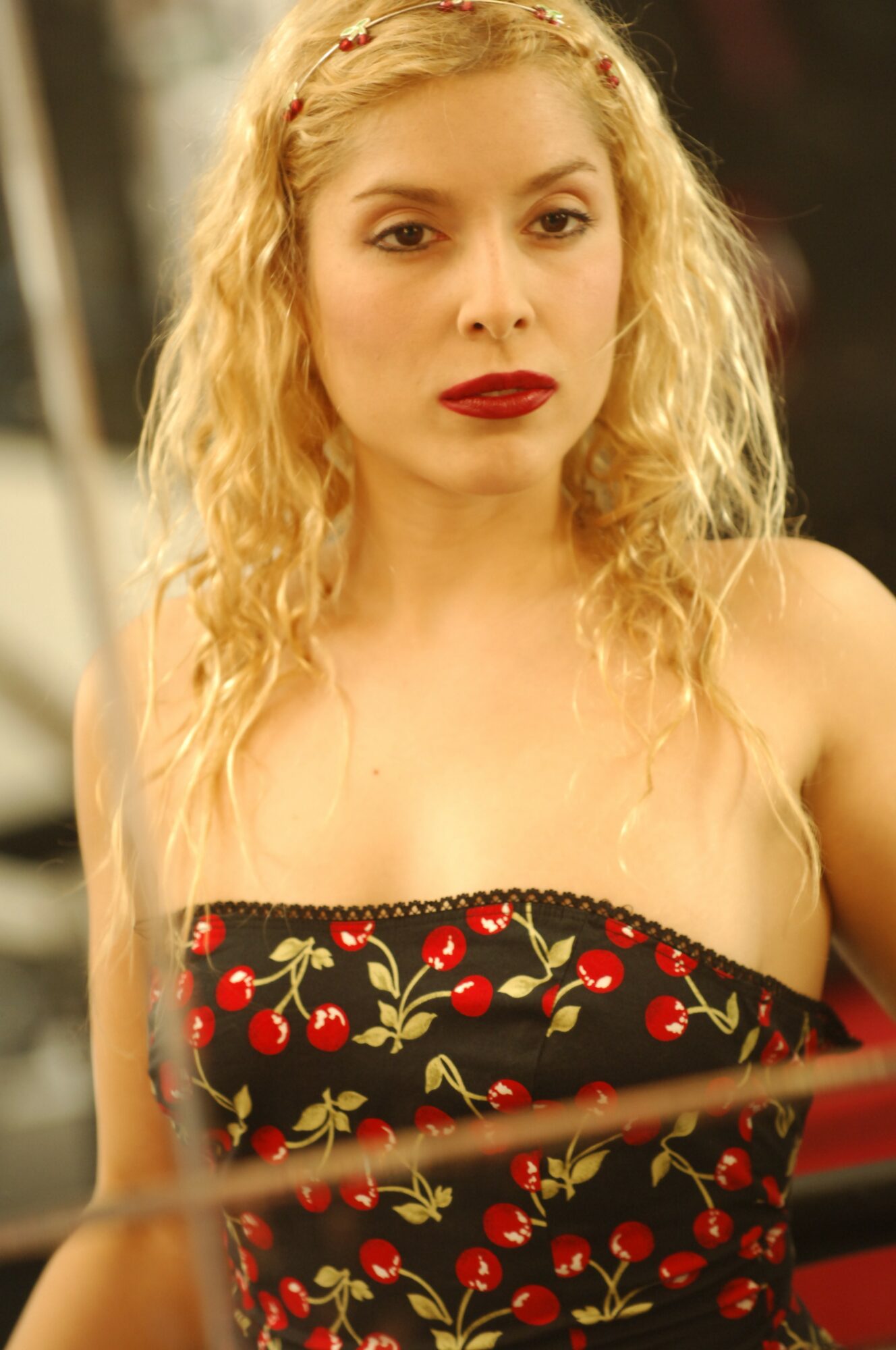 Young woman with curly blonde hair and red lipstick wearing a cherry-patterned strapless top.